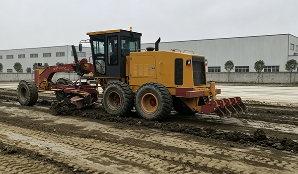 A yellow motor grader equipped with a rear ripper pulls a road recycler, working on muddy construction ground marked with tire tracks. Modern white factory buildings stand in the background under overcast daylight. The real-site scene demonstrates the powerful working performance of the construction machinery with attachments. A yellow motor grader equipped with a rear ripper pulls a road recycler, working on muddy construction ground marked with tire tracks. Modern white factory buildings stand in the background under overcast daylight. The real-site scene demonstrates the powerful working performance of the construction machinery with attachments.